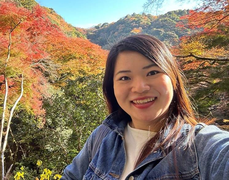 Woman smiling outdoors with colorful autumn trees and mountains in the background.